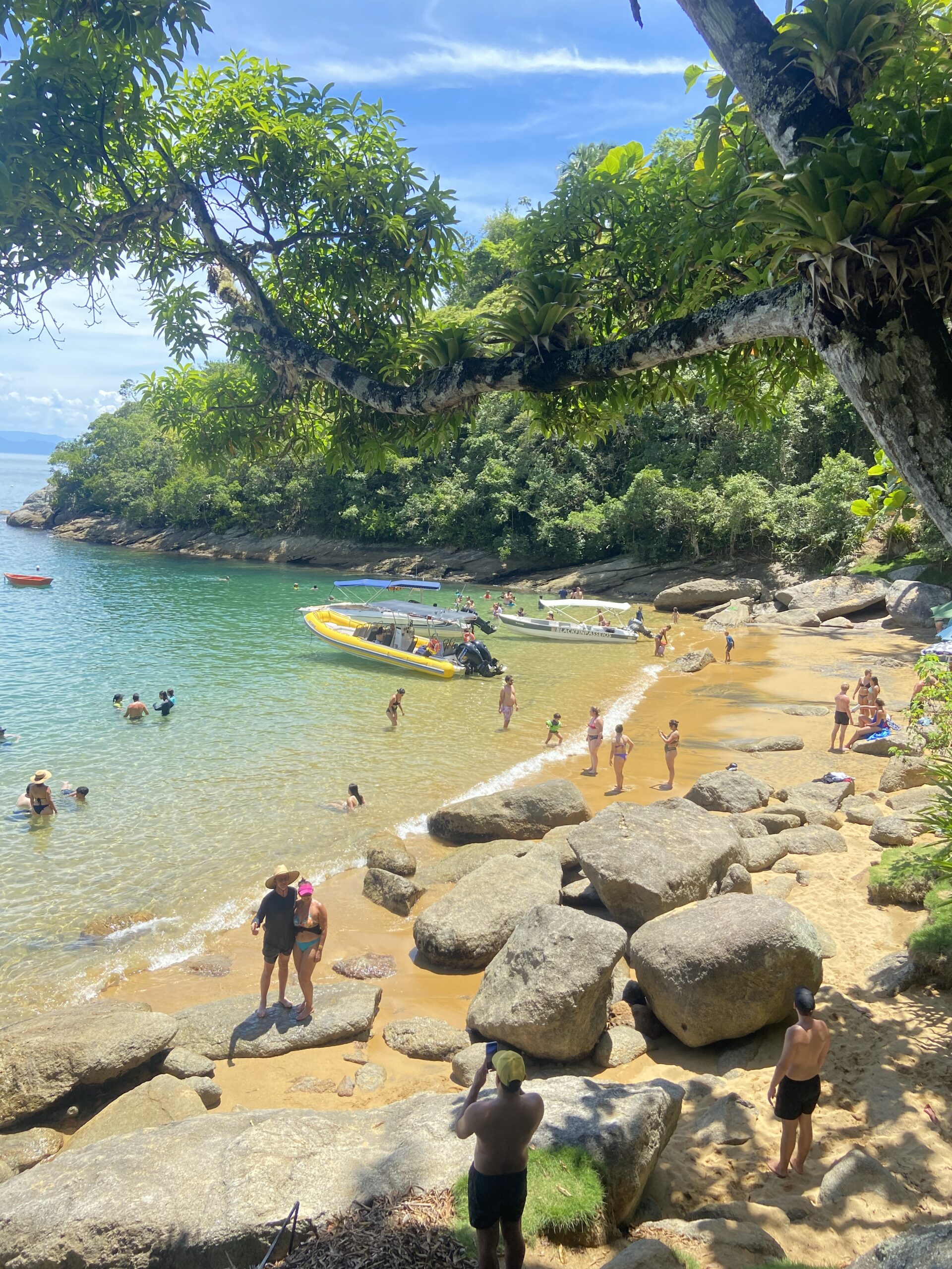 Enseada vista entre galhos e rochas, com pessoas nadando e caminhando na praia de areia dourada, várias embarcações — incluindo uma lancha inflável amarela — atracadas em águas verde-turquesa sob céu claro.