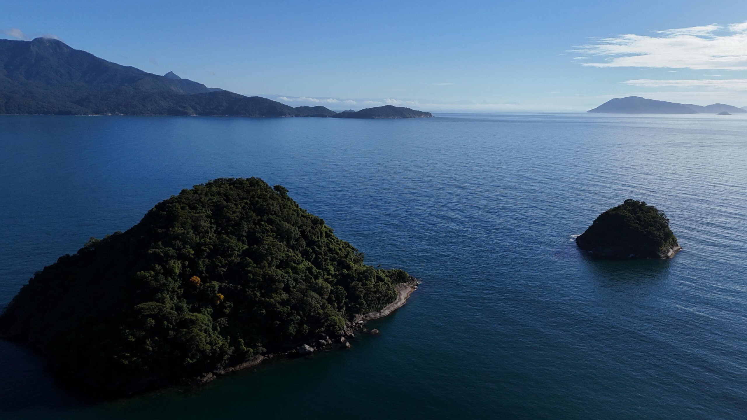 Duas pequenas ilhas cobertas por densa vegetação em mar calmo azul-escuro, com montanhas distantes sob céu claro no horizonte.
