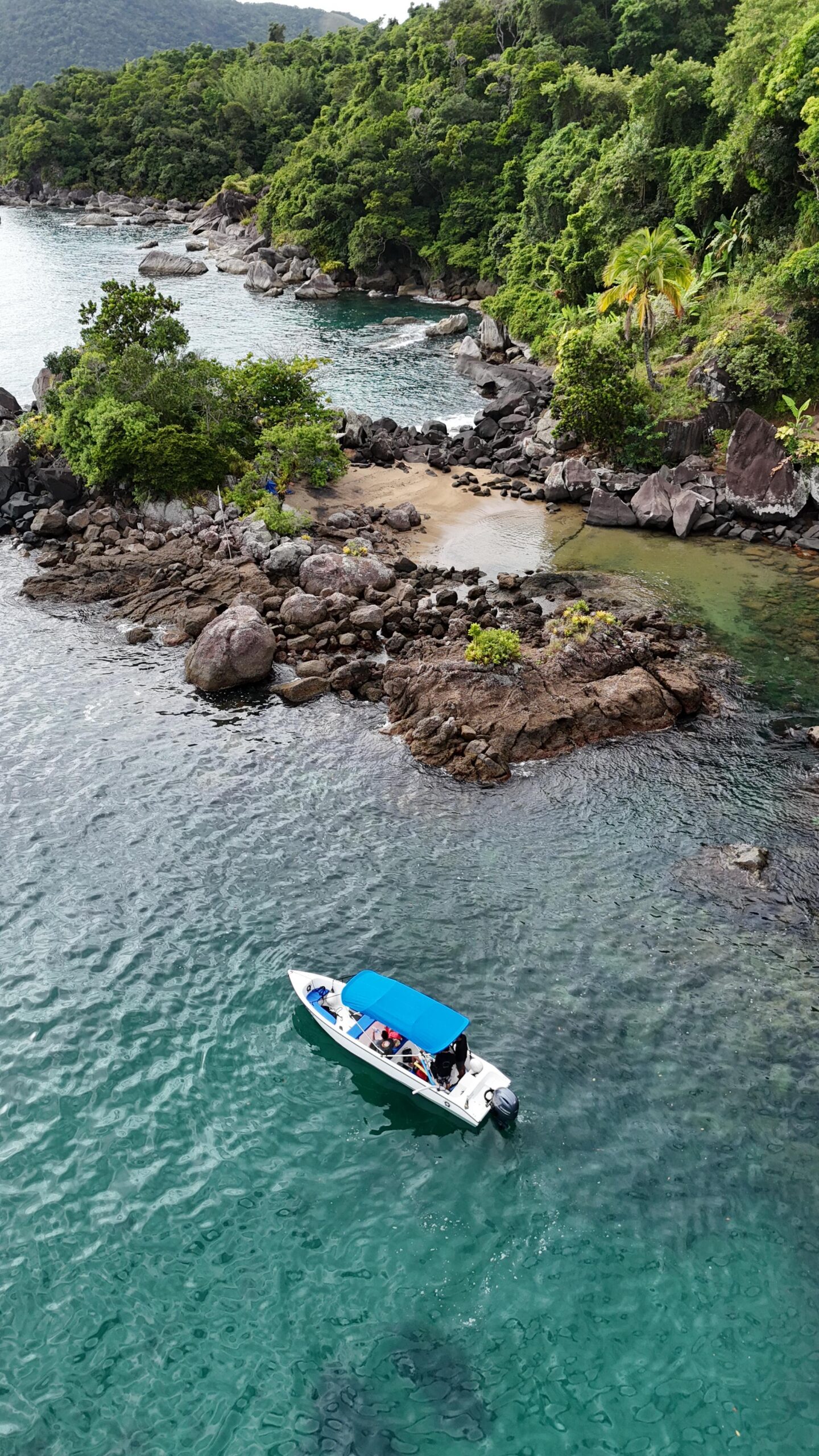 Pequeno barco branco com toldo azul flutuando em águas translúcidas verde-esmeralda próximas a uma costa rochosa, onde se vê mata densa cobrindo morros e uma pequena enseada arenosa.