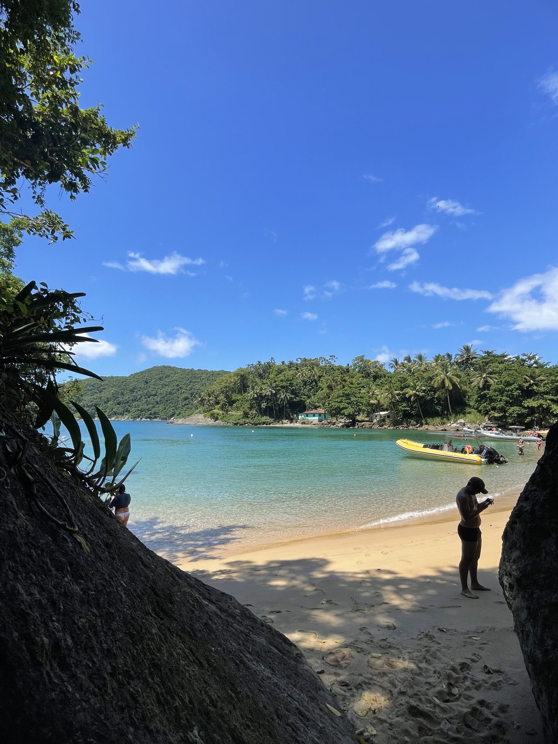 Enseada de areia dourada enquadrada por rochas e folhagens em primeiro plano, com águas cristalinas verde-turquesa, morro coberto de mata ao fundo, céu azul e, à direita, um homem em pé na areia conferindo o celular ao lado de uma lancha inflável amarela ancorada.