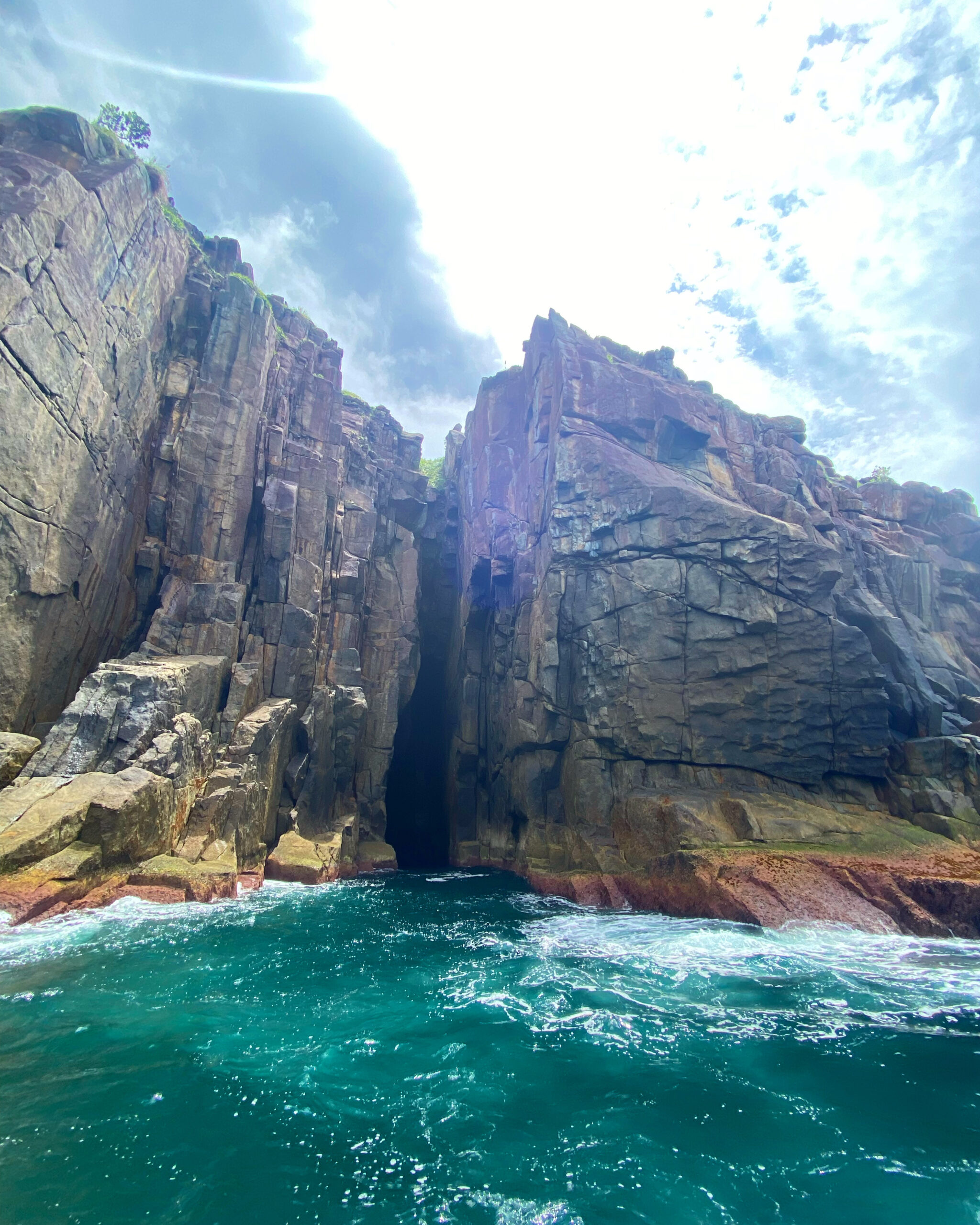 Alto paredão rochoso com fenda escura no centro e mar verde-esmeralda agitado na base, sob um céu parcialmente nublado.