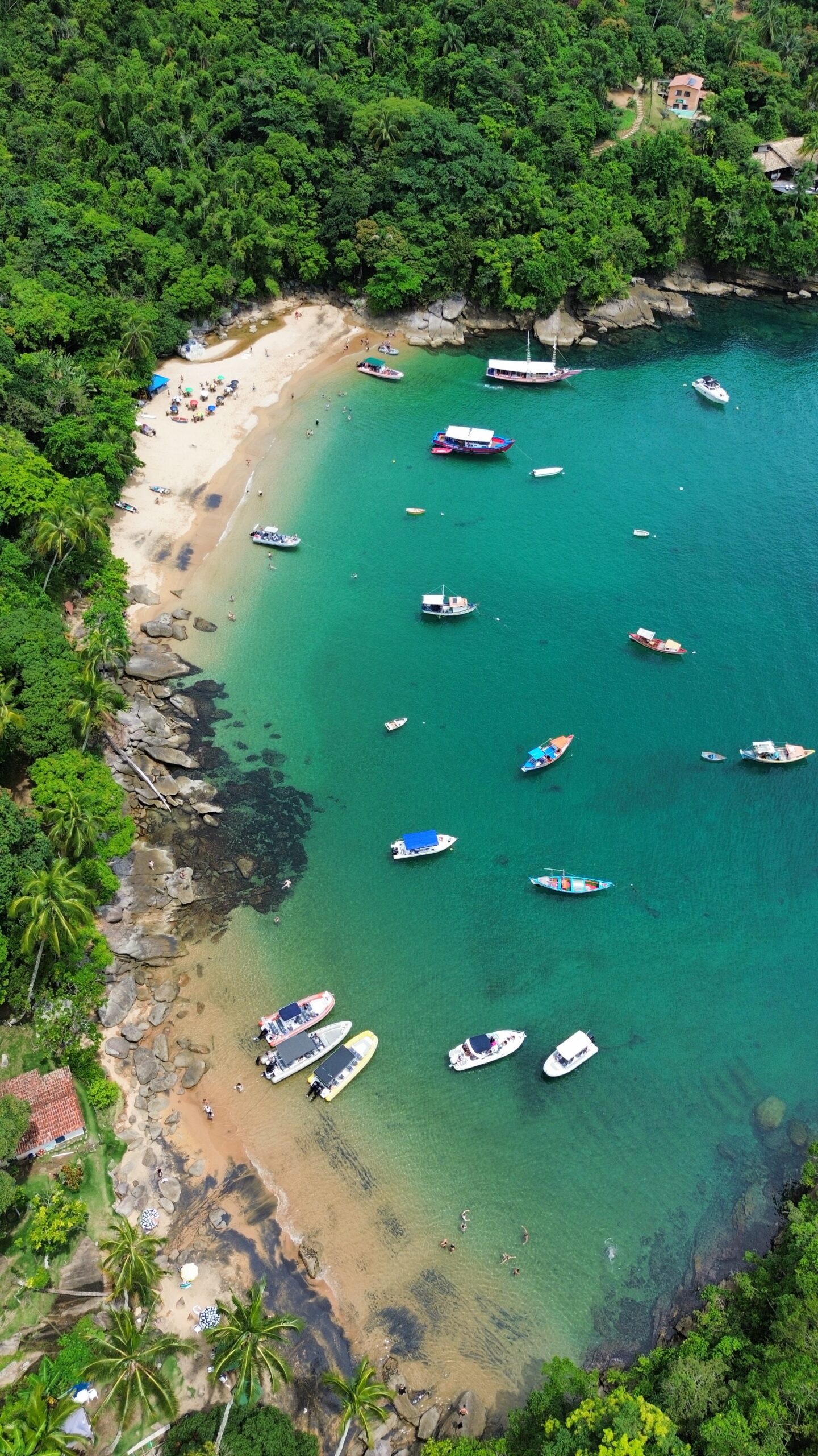 Pequena enseada de areia clara cercada por mata tropical densa, com diversos barcos ancorados em águas translúcidas verde-turquesa e pessoas na praia.