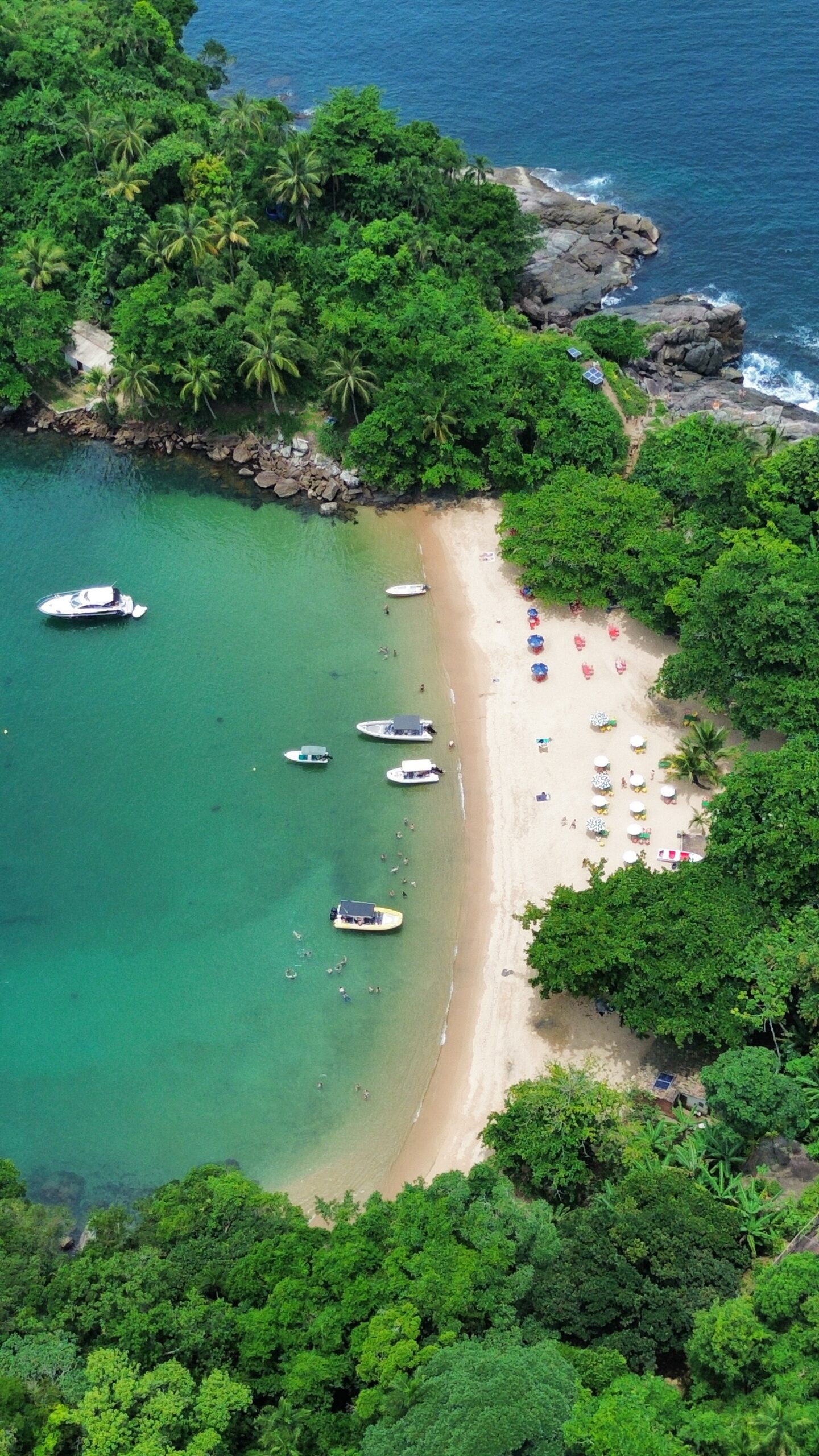 Vista aérea de praia de areias douradas em curva, ladeada por vegetação intensa, com guarda-sóis coloridos, banhistas e vários barcos em águas rasas.