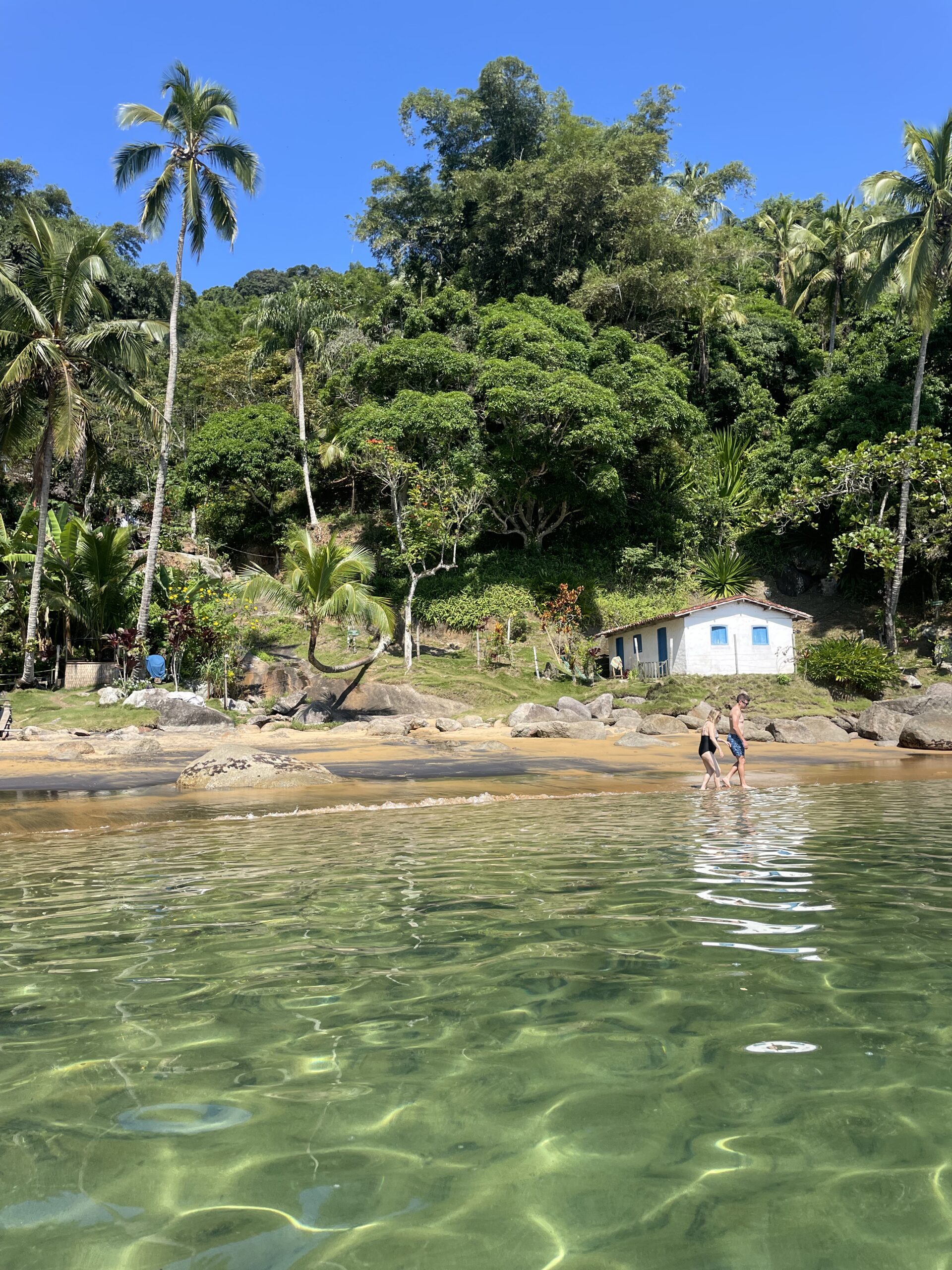 Águas rasas e transparentes refletindo a luz em primeiro plano, com duas pessoas caminhando na beira da praia arenosa diante de morro denso de coqueiros e pequena casa branca de janelas azuis à beira da mata.