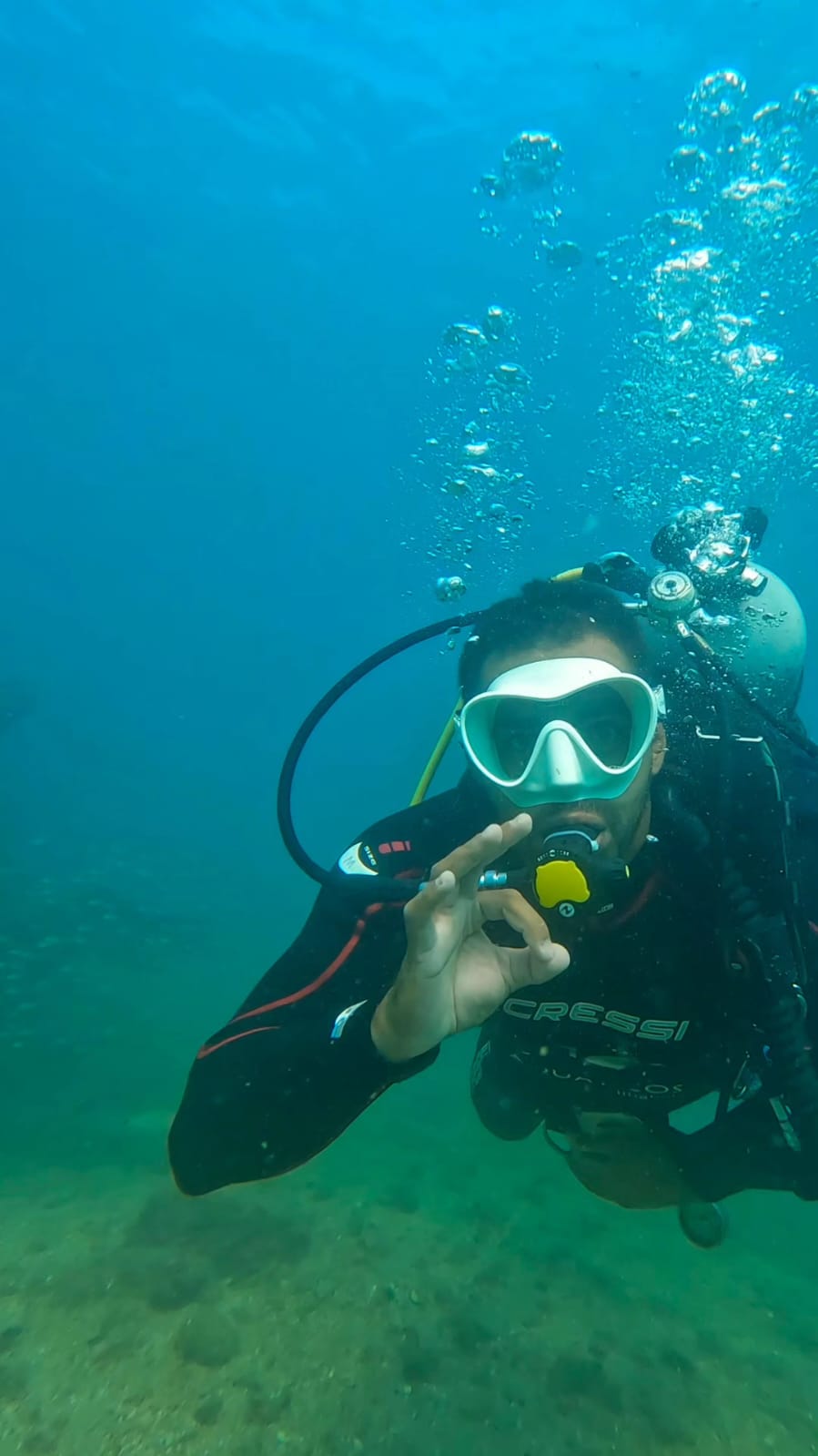 Mergulhador subaquático com máscara branca e cilindro de ar, fazendo o sinal de 'ok' com a mão enquanto bolhas sobem em volta.